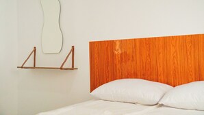 Une chambre à coucher avec une tête de lit en bois, un lit blanc et une étagère en bois surmontée d’un miroir.