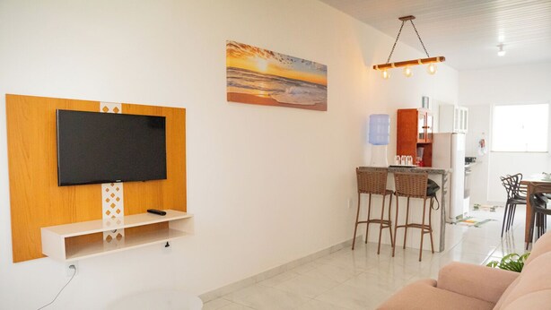 Un salon moderne avec un téléviseur à écran plat fixé sur un panneau en bois, une petite table blanche et un canapé beige. On aperçoit un coin repas avec deux chaises en osier et, au fond, une cuisine.