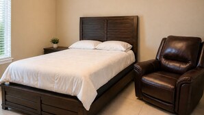 Een slaapkamer met een bed, nachtkastje, potplant en een lederen fauteuil.