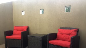 Twee rieten stoelen met rode kussens en een klein tafeltje, geplaatst tegen een structuurrijke muur met drie kleine ramen.