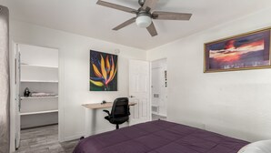 A bedroom with a bed, a desk, a chair, a ceiling fan, and a painting on the wall.