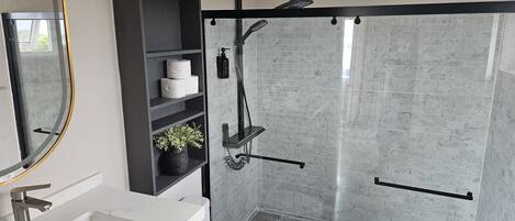Une salle de bain moderne dotée d’une douche à parois de verre, d’un lavabo blanc, de toilettes et d’un meuble de rangement noir avec des étagères.