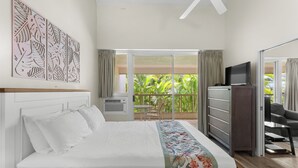 A hotel room with a bed, a TV, a desk, and a balcony with a view of greenery. - Maui Banyan A-202 by Maui Rental Group (Kihei)
