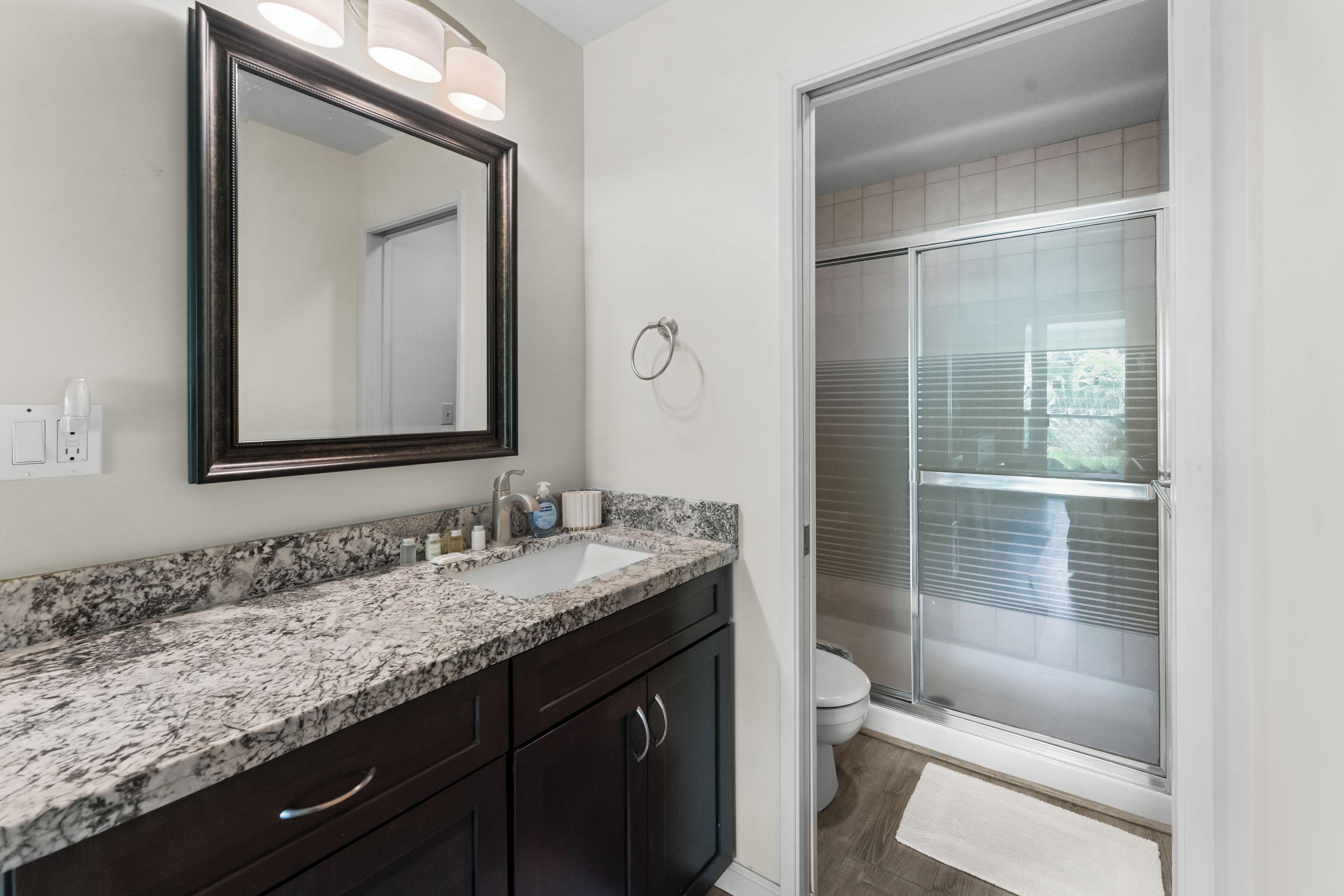 A bathroom with a granite countertop, dark cabinets, a large mirror, and a glass shower enclosure.