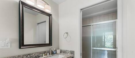 A bathroom with a granite countertop, dark cabinets, a large mirror, and a glass shower enclosure.