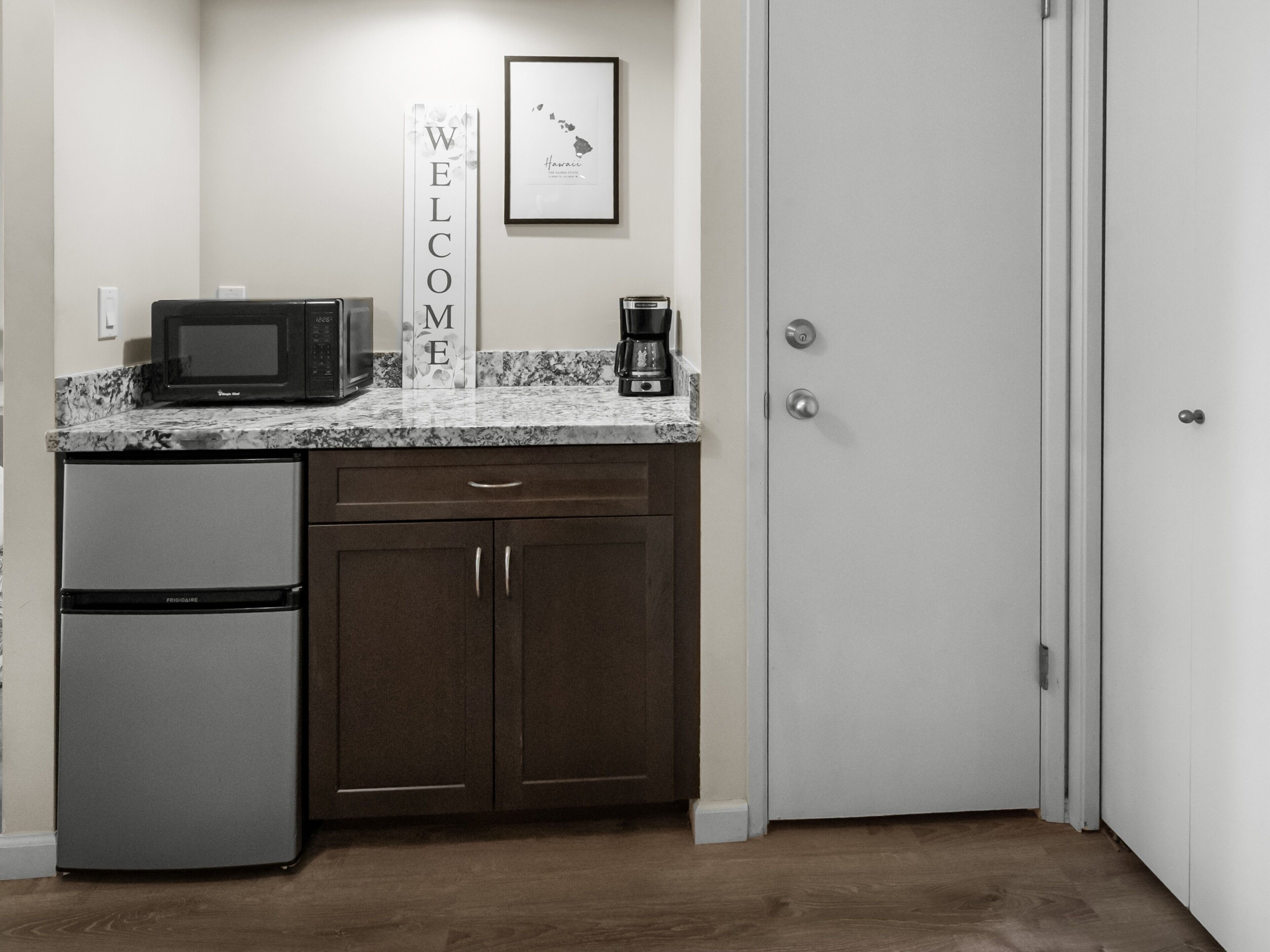 A compact kitchen with a refrigerator, microwave, coffee maker, and a welcome sign.