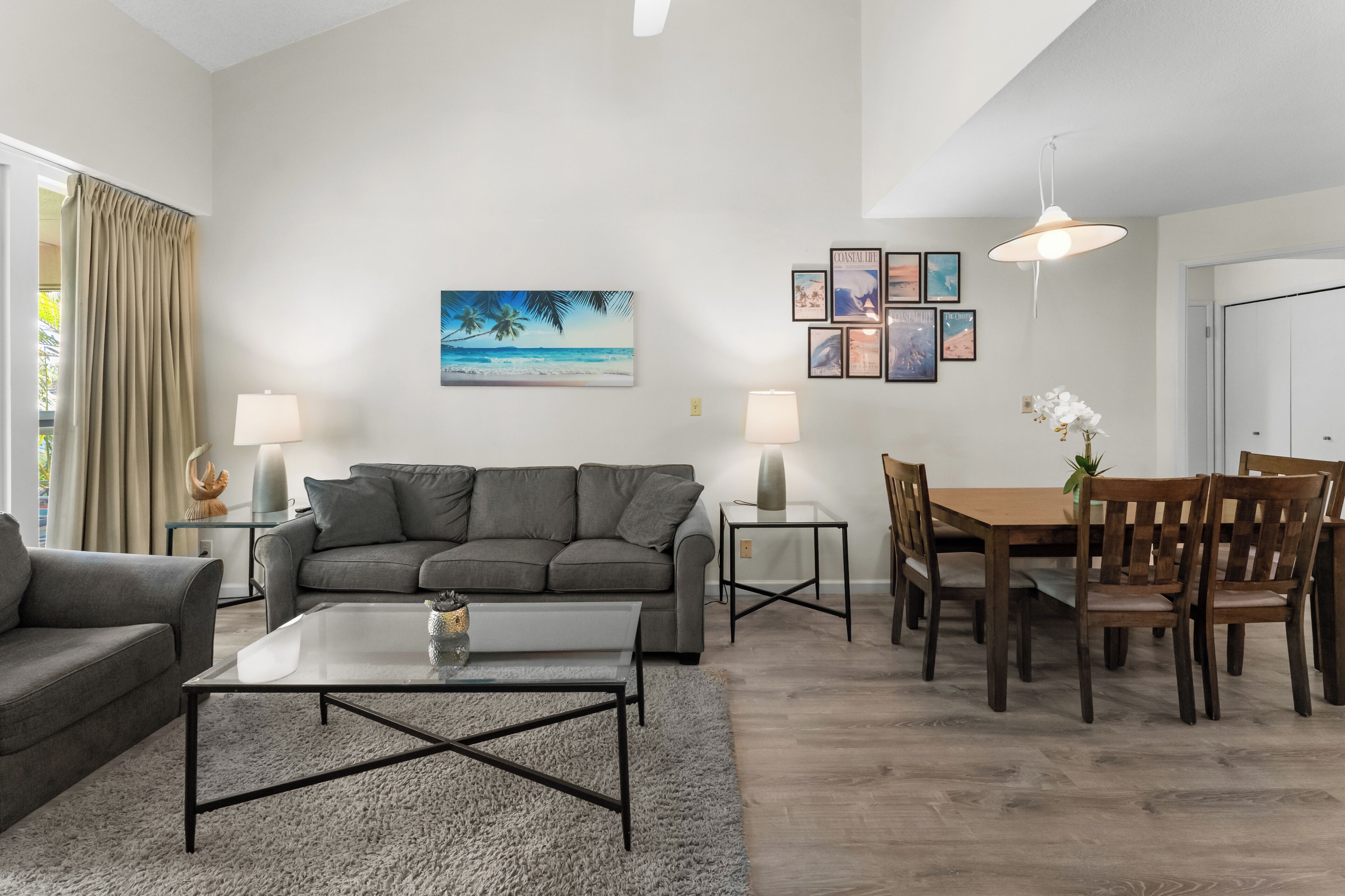 Ein modernes Wohnzimmer mit grauem Sofa, Couchtisch und Essbereich mit Holzsesseln und Tisch. An den Wänden befinden sich Bilderrahmen und eine Pendelleuchte.