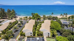 A coastal residential area with a beach view, featuring multiple-story buildings, parking spaces, and lush greenery. - Maui Banyan A-202 by Maui Rental Group (Kihei)