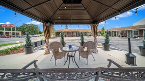 An outdoor covered patio with wicker chairs and a round table, potted plants, and a view of a street with buildings in the background.