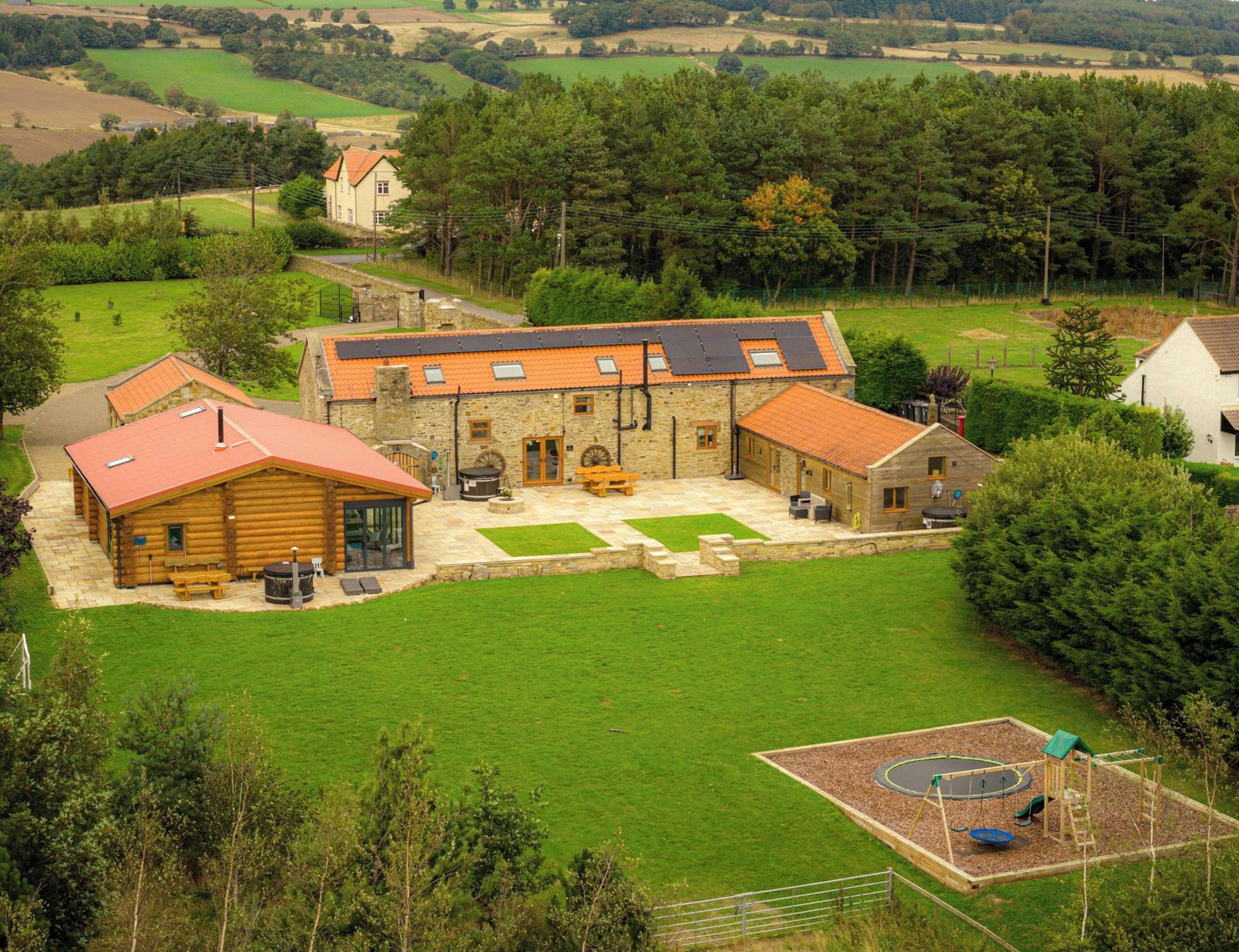 Un bâtiment en pierre avec un toit rouge, un chalet en bois et une aire de jeux avec des balançoires et un trampoline.