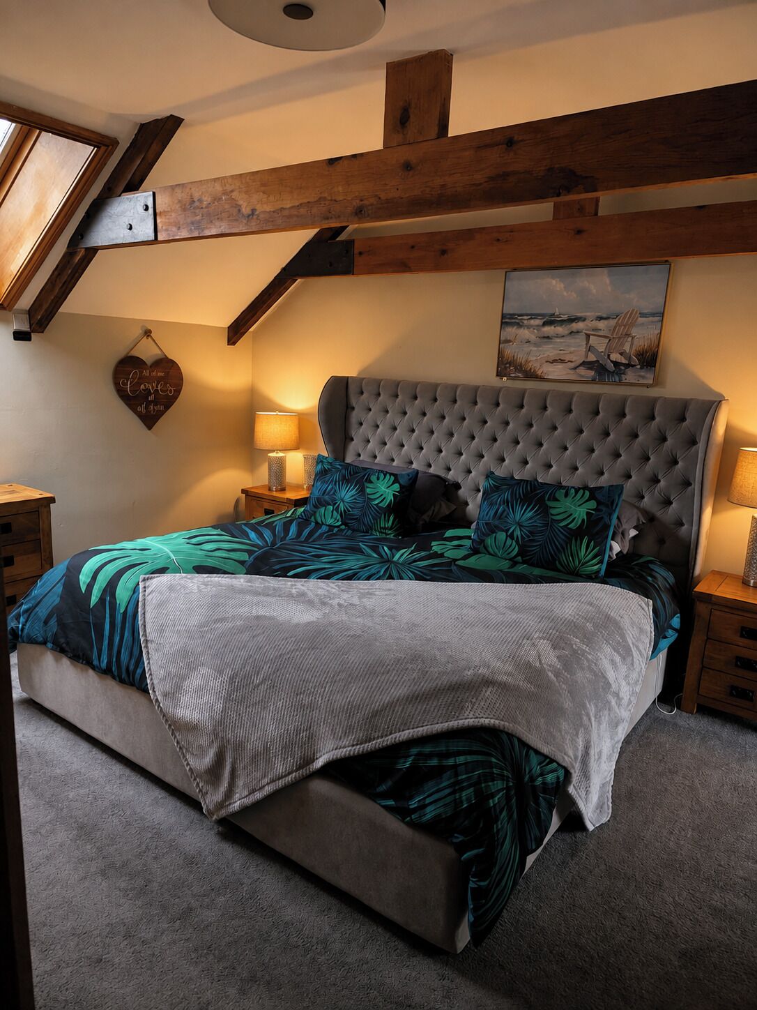 Un dormitorio con cabecera capitonada, cama con colcha estampada de hojas, decoración de pared con forma de corazón y cuadro de una escena de playa.