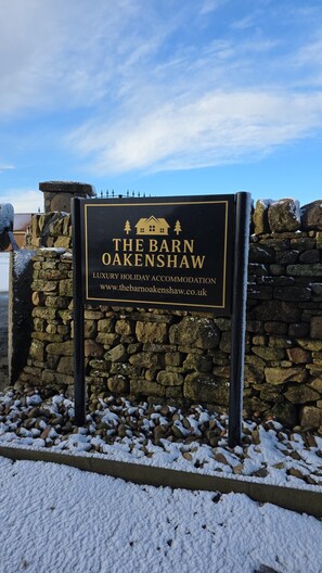 A sign for The Barn Oakenshaw, a luxury holiday accommodation, with a stone wall and snow-covered ground.