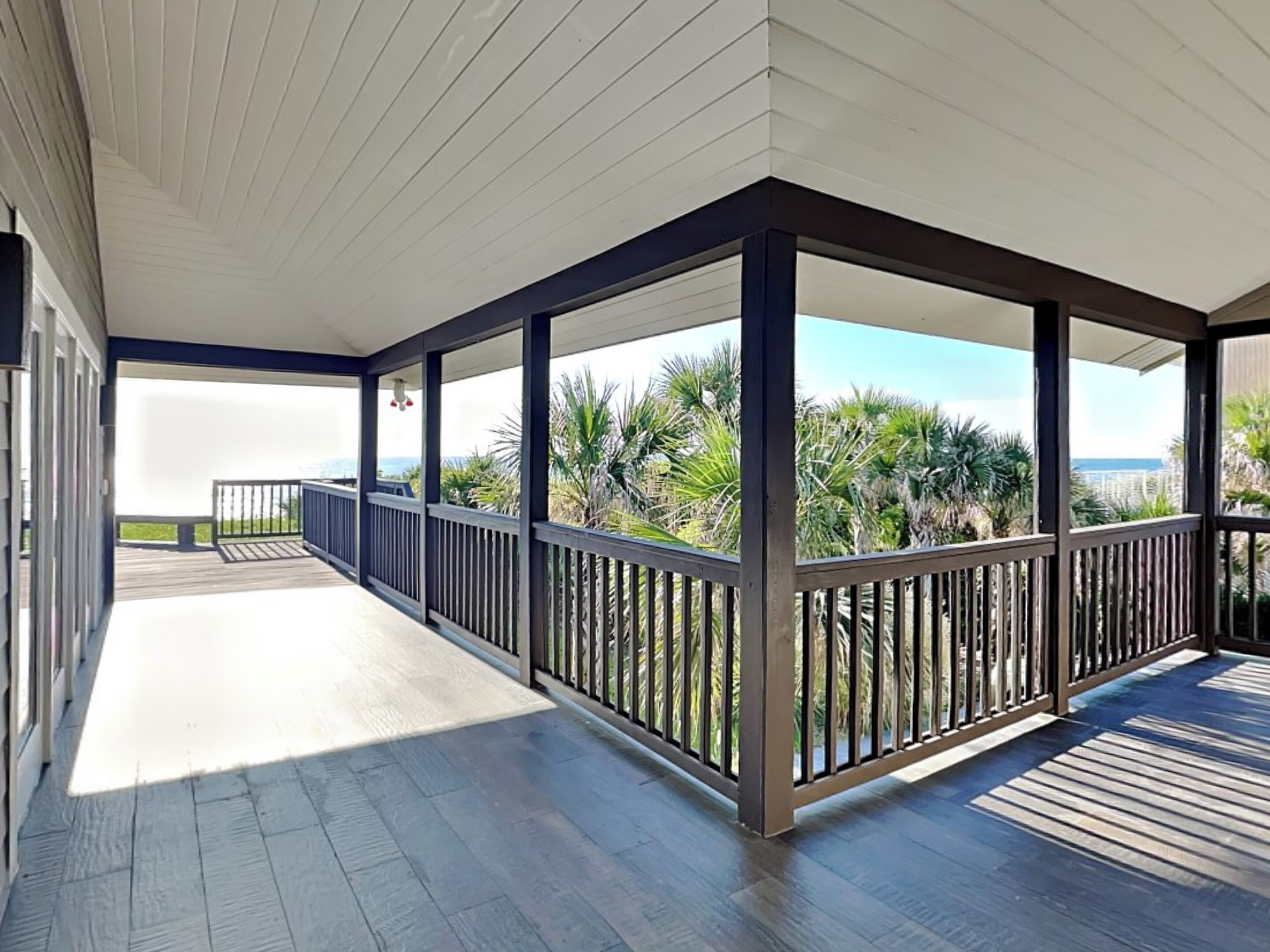 A covered outdoor area with a wooden deck, metal railings, and a view of palm trees and the ocean.