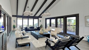 A spacious living room with a dark leather sofa, white armchairs, and a black coffee table. Large windows provide a view of the outdoors. - Ocean Commotion by Distinctive Beach Rentals (Ponte Vedra Beach)
