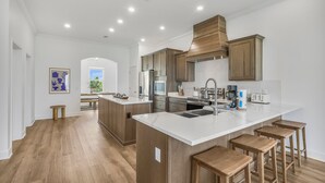 A modern kitchen with white countertops, wooden cabinets, and a central island with stools. - New! 5BD+Private Pool! 3 Decks with amazing views! (Gulf Shores)