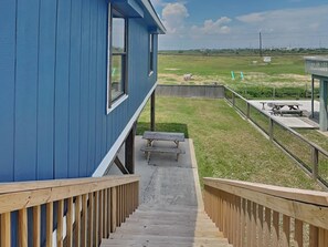 A blue building with a wooden deck leading to a grassy area with a picnic table and a small structure. - Ocean Oasis by Ryson Vacation Rentals (Galveston)