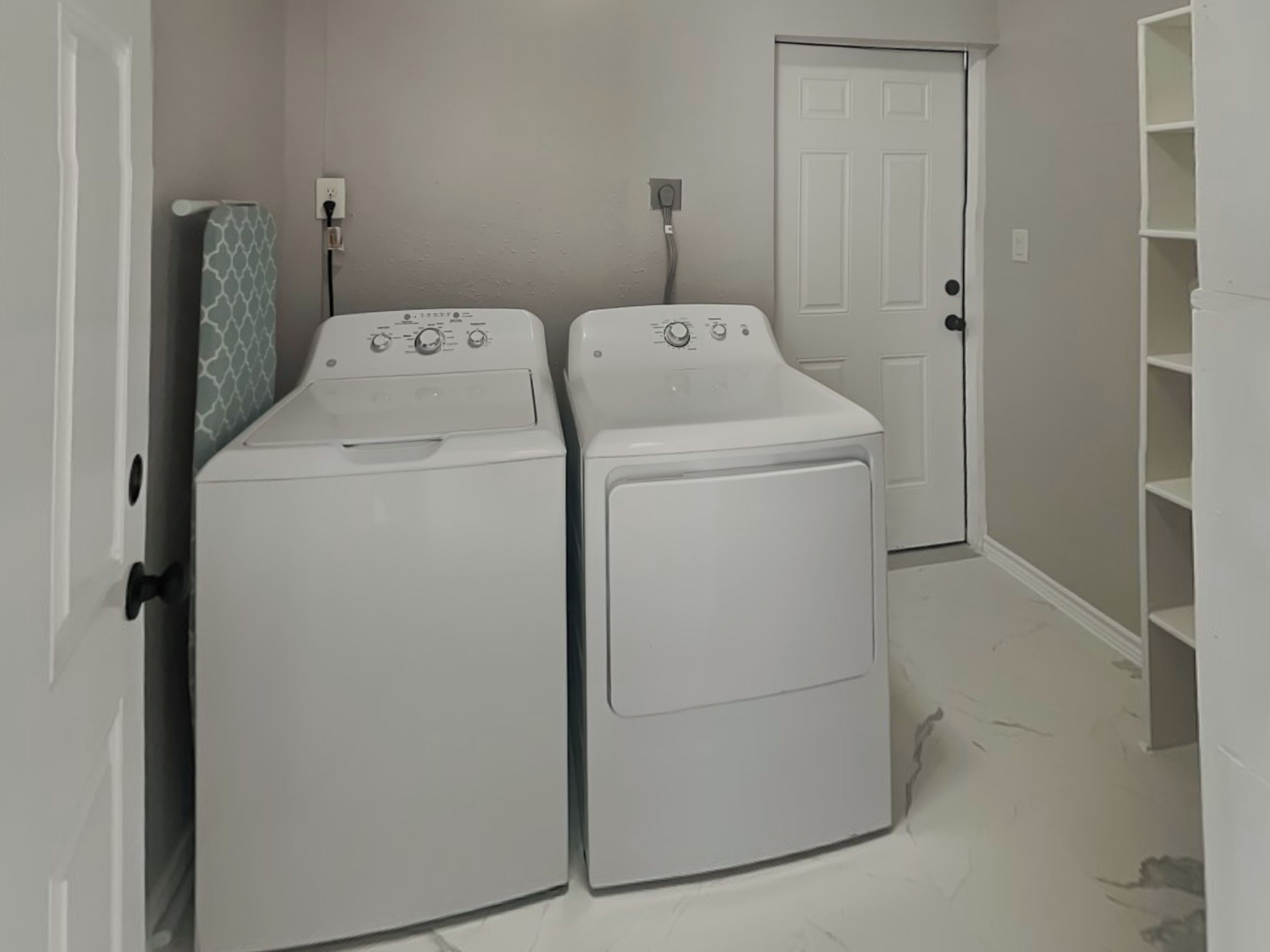 A laundry room with two front-loading washing machines, a door, and a shelving unit.