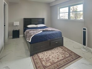 A bedroom with a bed, a nightstand, a lamp, a window, and a radiator. - Ocean Oasis by Ryson Vacation Rentals (Galveston)