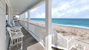 A covered porch with white railings and wicker chairs, overlooking a beach with a wooden walkway leading to the shore. - Ocean Paradise by Distinctive Beach Rentals (Ponte Vedra Beach)