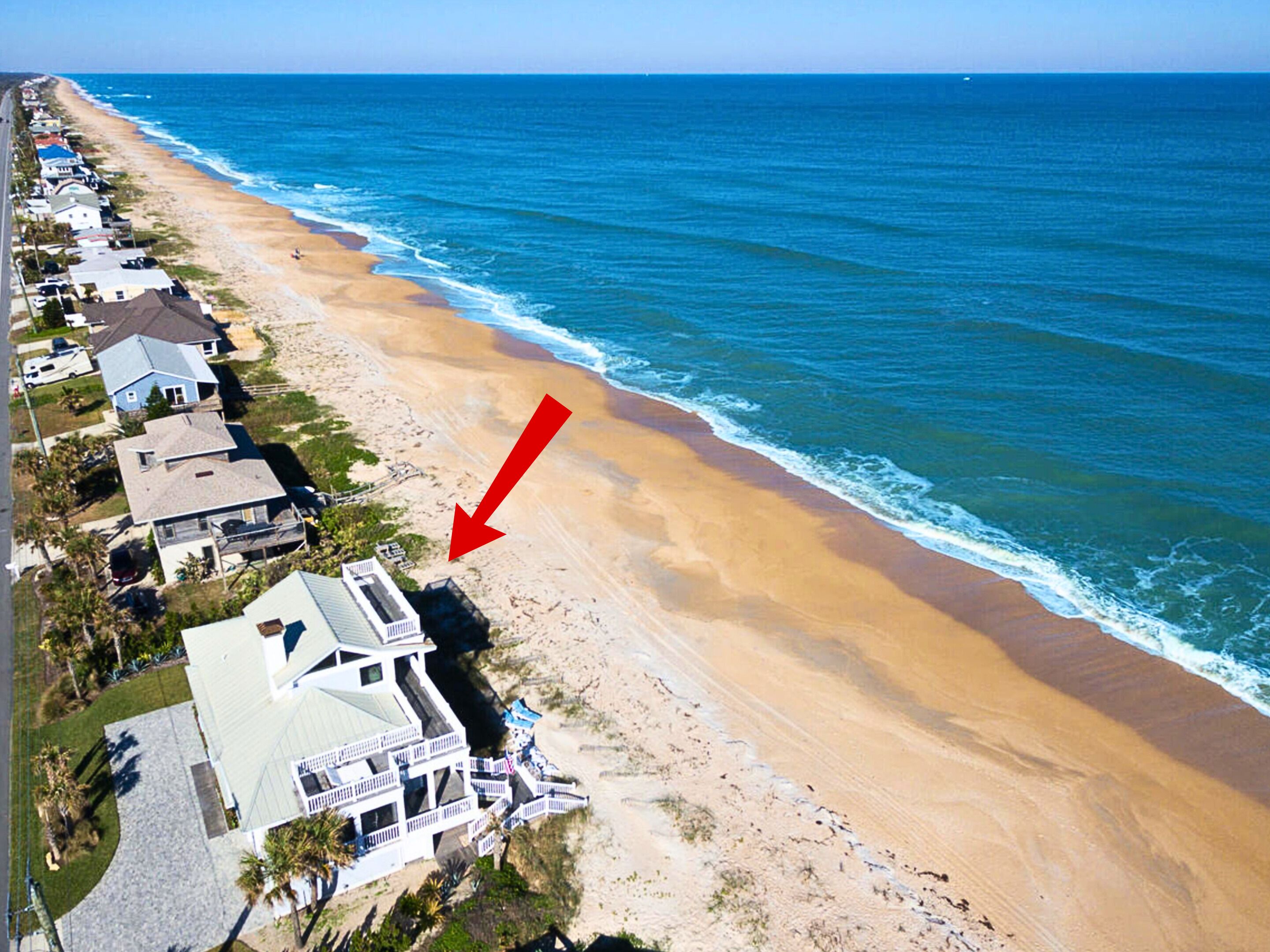 An aerial view of a coastal area with a row of houses along the beach, a large building with a white facade and multiple balconies, and a red arrow pointing to a specific location.