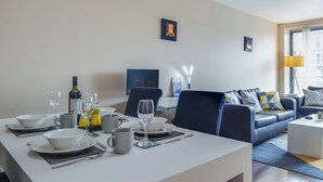 Un comedor moderno con una mesa blanca dispuesta para dos comensales, un sofá azul oscuro y vista a la ciudad a través de un ventanal grande.
