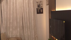 A living room with a sofa, a flat-screen TV, a decorative wall piece, and a curtain. - Your Cozy 1-bedroom, for short or long stays; minutes from the ocean and dining! (Dar es Salaam)