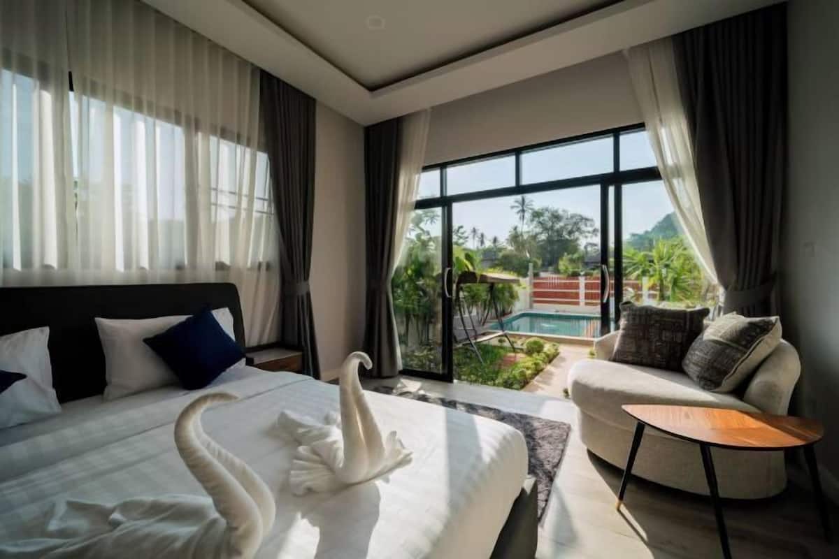 A hotel room with a bed, a sofa, a small table, and a view of a pool and greenery.