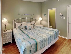 A bedroom with a bed, two bedside tables, a nightstand, a wall-mounted lamp, and a door. - Sea Turtle by Ryson Vacation Rentals (Galveston)
