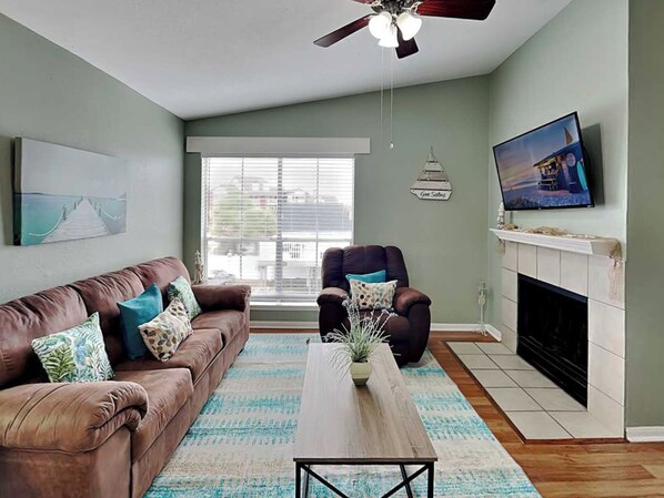 A living room with a brown leather sofa, a wooden coffee table, a fireplace, a television, and a ceiling fan. - Sea Turtle by Ryson Vacation Rentals (Galveston)