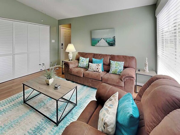A living room with a brown sofa, a coffee table, a lamp, and a painting on the wall. - Sea Turtle by Ryson Vacation Rentals (Galveston)