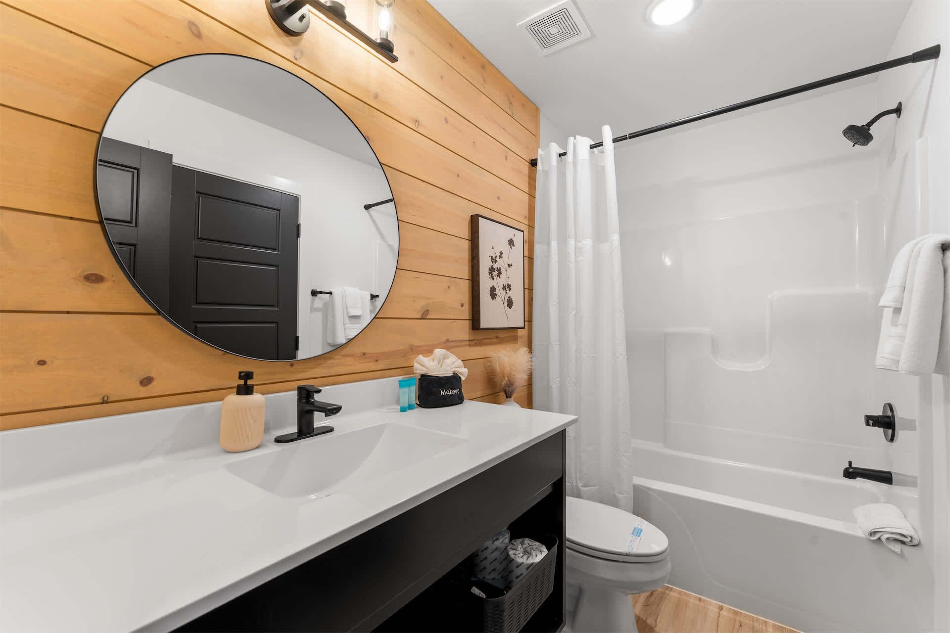 A modern bathroom with a wooden vanity, a round mirror, a toilet, and a shower area.