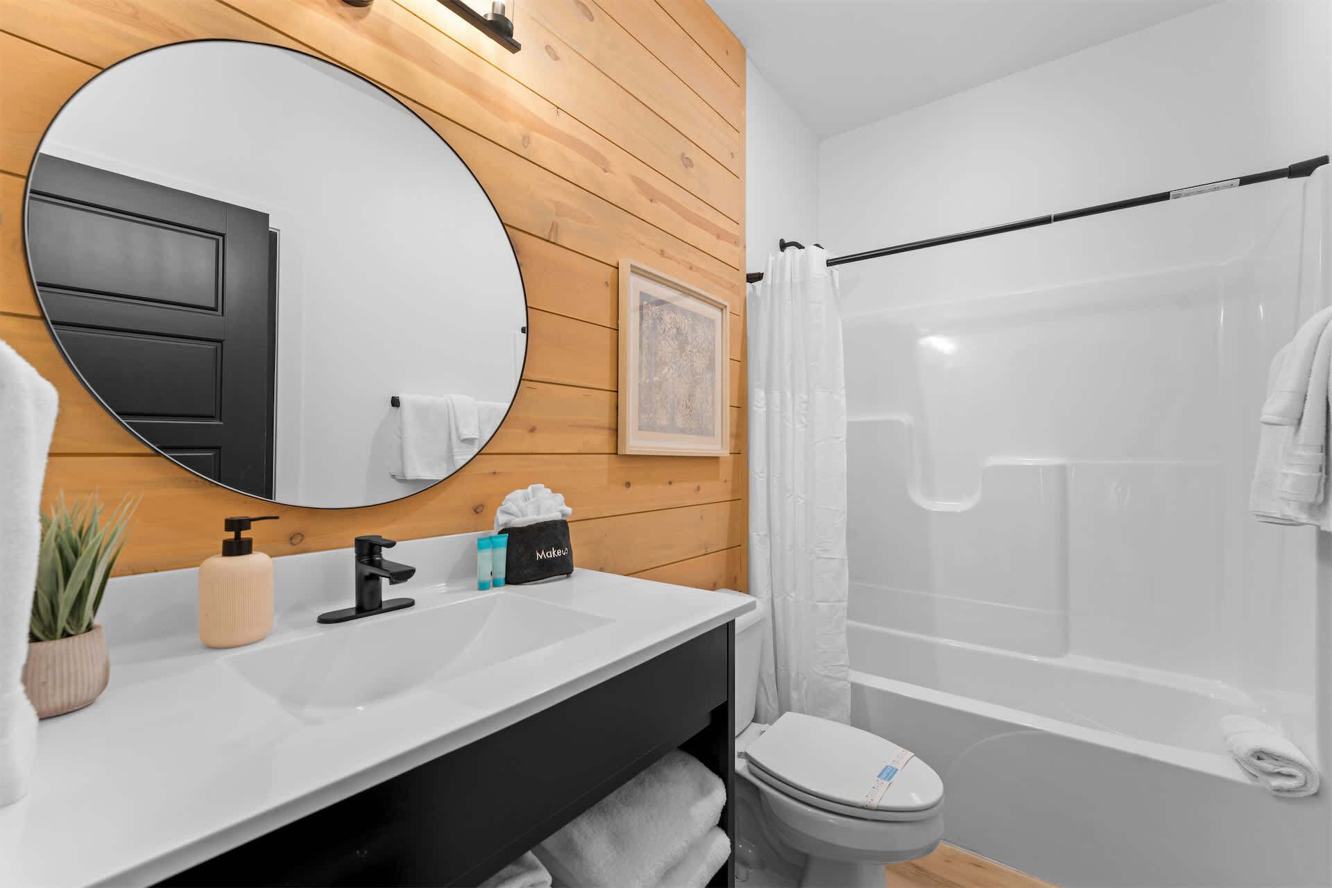 Ein modernes Badezimmer mit einem runden Spiegel, Wandverkleidung aus Holz, einem weißen Waschbecken, einem schwarzen Wasserhahn, einer Toilette und einer Duschkabine aus Glas.