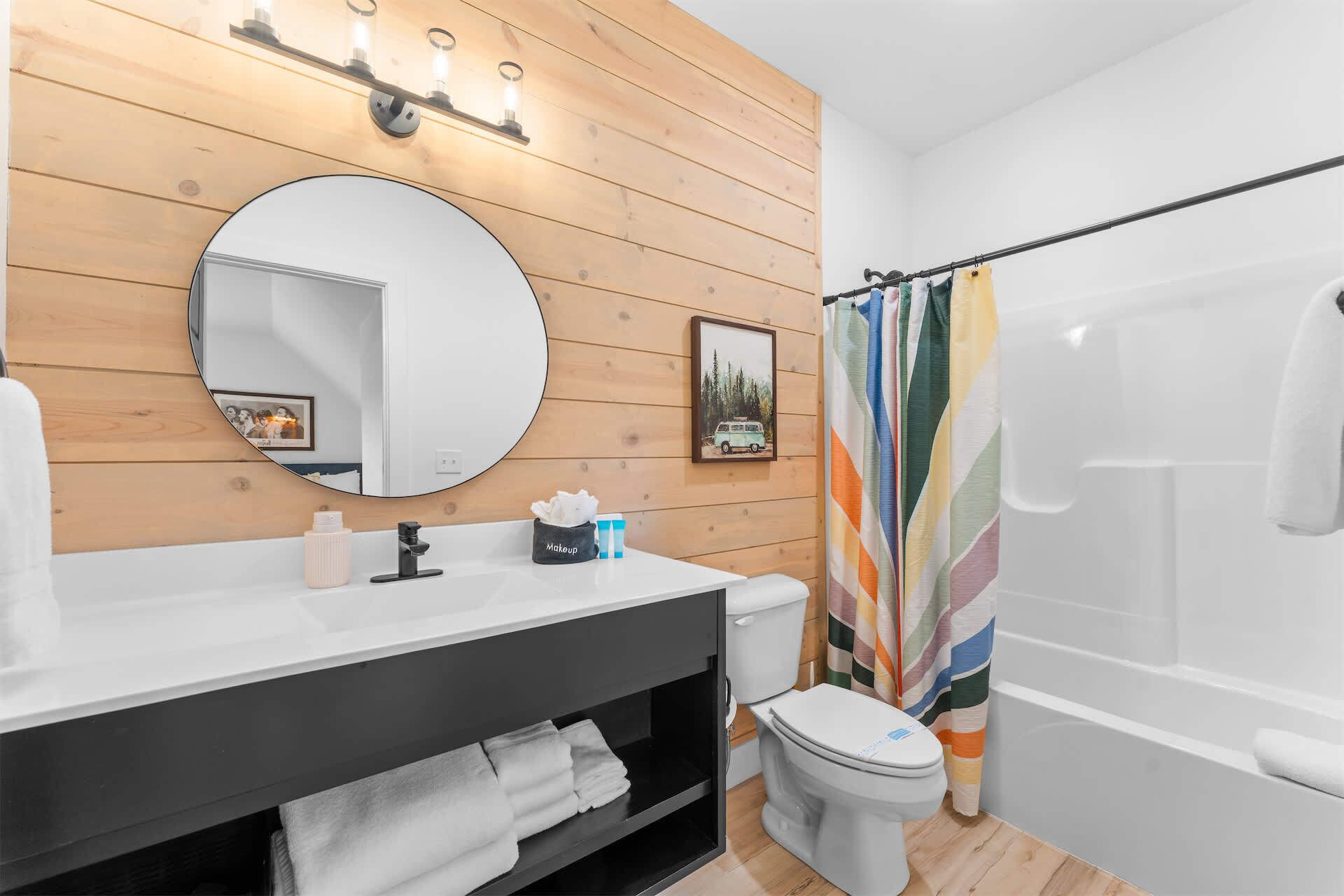 A bathroom with a wooden vanity, a round mirror, a toilet, and a shower with a colorful striped curtain.