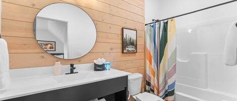 A bathroom with a wooden vanity, a round mirror, a toilet, and a shower with a colorful striped curtain.