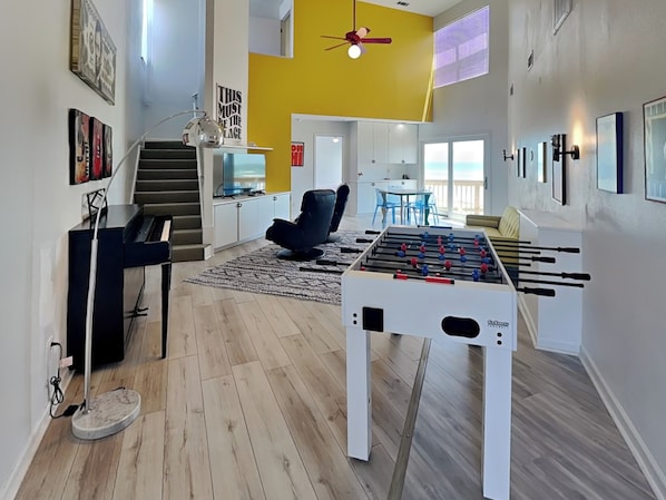 A modern living room with a foosball table, a piano, and a staircase leading to a balcony. - Pirates Beach Retreat by Ryson Vacation Rentals (Galveston)