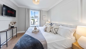 A hotel room with a bed, a TV mounted on the wall, a bedside table, and a window with curtains. - Long Stay Ready | King Bed | Kitchen | WiFi | Town (Kent)