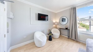 A modern living room with a flat-screen TV mounted on the wall, two white armchairs, a small side table with a vase, and a view of the outside through a large window. - Modern King Bed WiFi Kitchen Perfect for Long Stay (Kent)