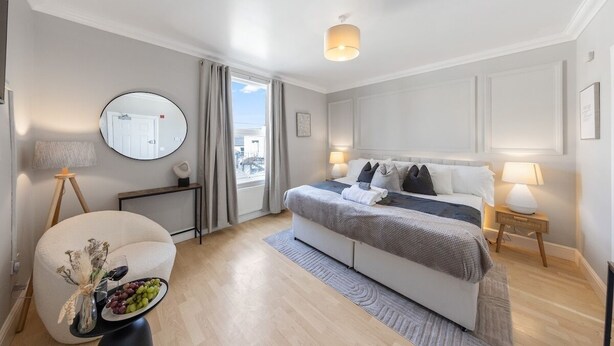 A modern hotel room with a large bed, bedside lamps, a round mirror, a small table with a vase and grapes, and a view of the outside through a window. - Modern King Bed WiFi Kitchen Perfect for Long Stay (Kent)