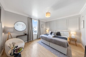 Ein modernes Hotelzimmer mit einem großen Bett, Nachttischlampen, einem runden Spiegel, einem kleinen Tisch mit einer Vase und Trauben sowie Blick durch ein Fenster nach draußen.