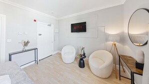 A modern living room with a flat-screen TV, a round mirror, a white armchair, a bean bag chair, a small side table with a vase, and a wooden side table. - Modern King Bed WiFi Kitchen Perfect for Long Stay (Kent)