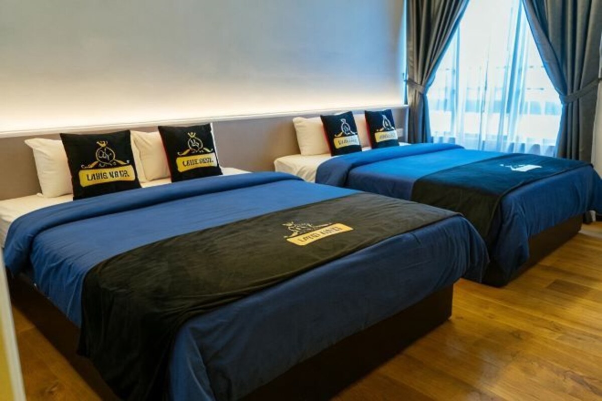 Two beds with blue bedding, pillows with logos, a wooden floor, and a window with curtains.