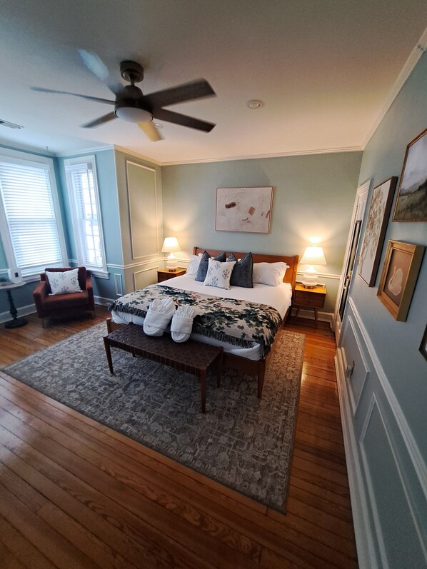 Une chambre à coucher comprenant un lit, deux tables de chevet, une chaise, un ventilateur de plafond et une grande fenêtre avec des stores.