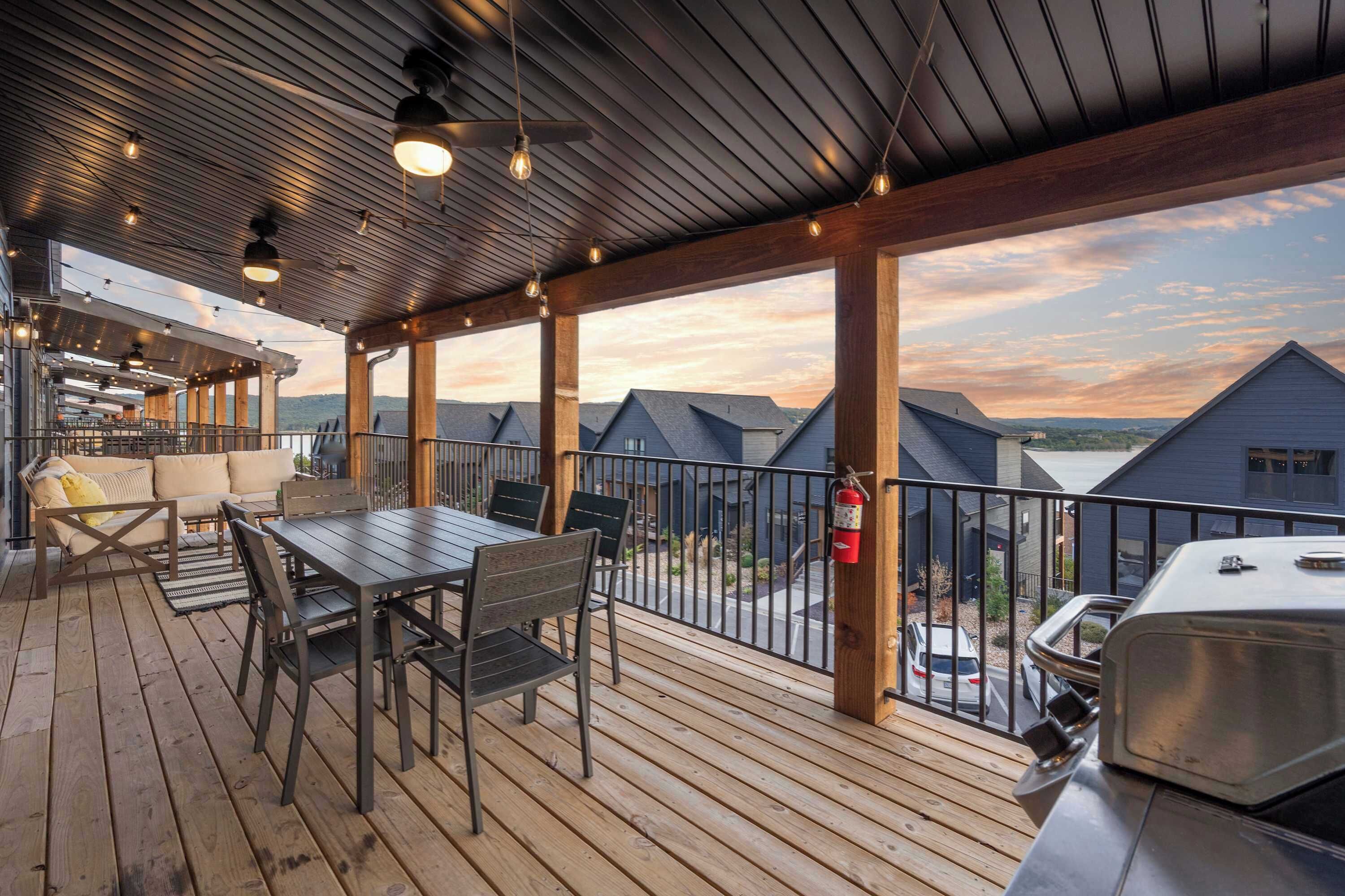 A covered outdoor deck with a dining table and chairs, a grill, and a view of neighboring houses.