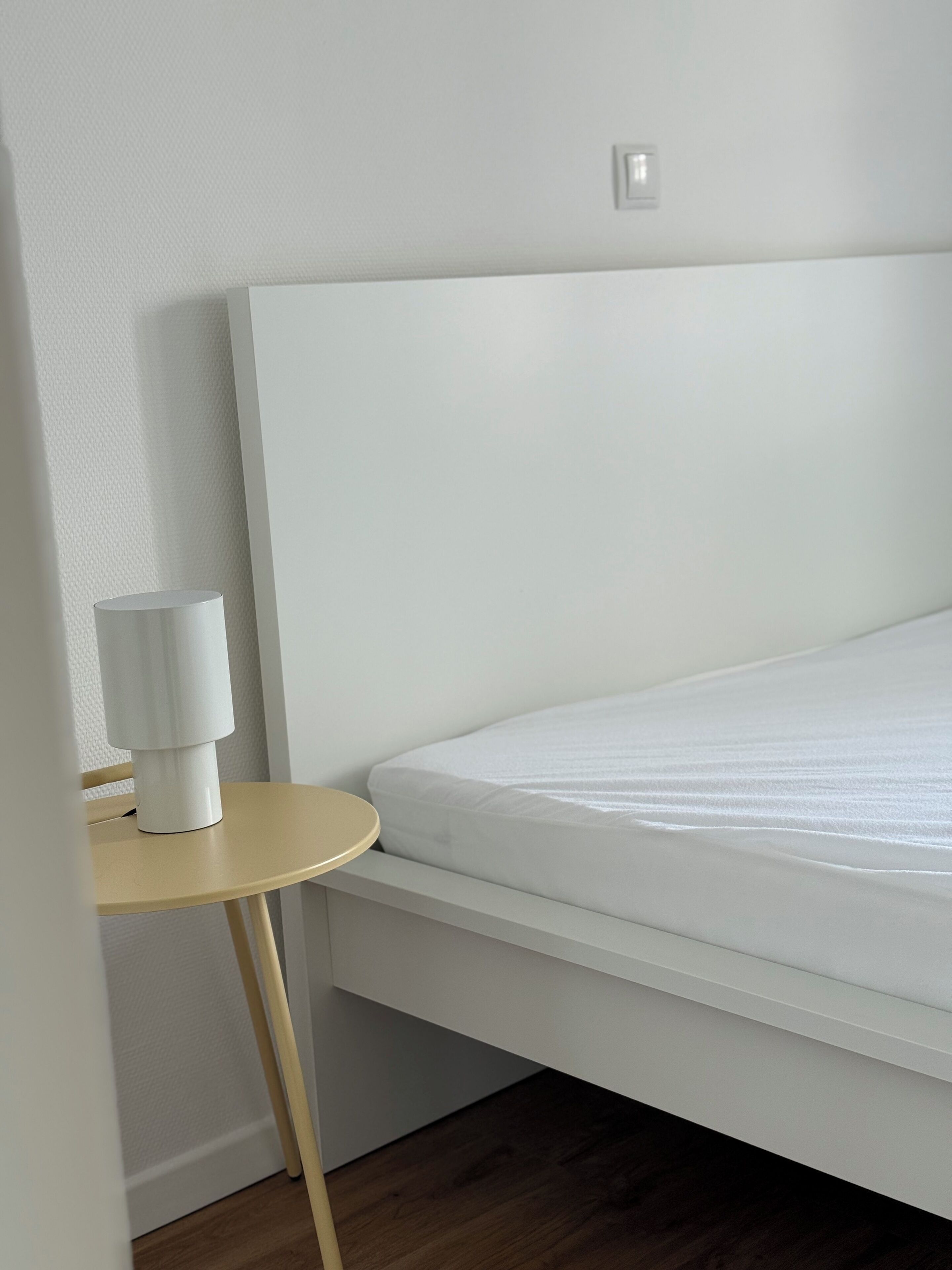 A neatly made bed with a white headboard, a small round table with a lamp, and a wooden floor.