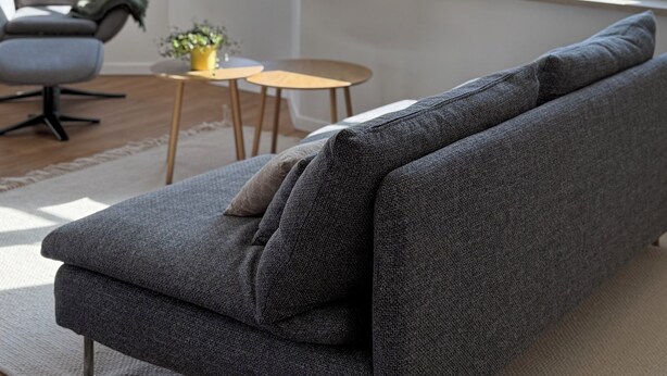 Una sala de estar moderna con un sofá gris, una mesa redonda de madera y una silla. Hay ventanas grandes con vistas al exterior.