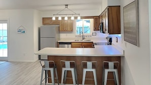A modern kitchen with a central island, bar stools, wooden cabinets, and a stainless steel refrigerator. - Rivers Edge Retreat by Distinctive Beach Rentals (St. Augustine)