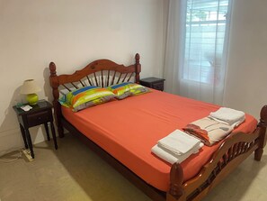 Una cama bien hecha con tablero de madera, una mesita de noche con lámpara y una ventana con persianas.
