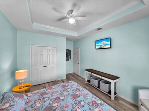 Un dormitorio con cama, ventilador de techo, mesita de noche con lámpara, cómoda con espejo y televisor en la pared.