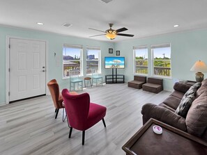 A spacious living room with a ceiling fan, a television, and a view of the outdoors through large windows. - Ibis Isle-Bolivar Peninsula, Sleeps 12, Fenced Side Yard, Walk to Beach, In Town (Bolivar Peninsula)