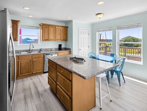 A kitchen with wooden cabinets, a granite countertop, and a dining area with blue chairs. - Ibis Isle-Bolivar Peninsula, Sleeps 12, Fenced Side Yard, Walk to Beach, In Town (Bolivar Peninsula)
